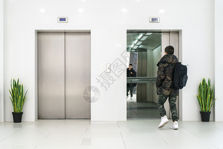 穿便衣和白色运动鞋的少年男孩进入电梯白色当代建筑内部花盆和白色墙中的花朵金属电梯门图片
