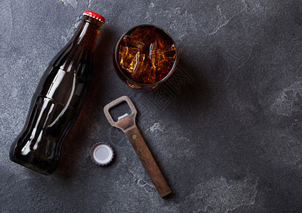 一瓶可乐苏打饮料和玻璃与冰块旁边的开瓶器在黑石上图片