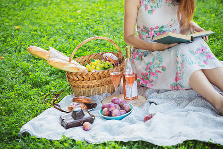 女孩坐在野餐篮子和面包葡萄酒眼镜和面包卷旁看书图片