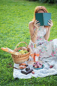 女孩坐在野餐篮子和面包葡萄酒眼镜和卷饼旁看书的女孩图片