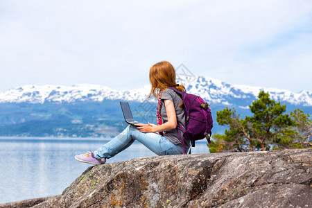 穿着笔记本电脑坐在山地和湖泊背景的岩石上图片