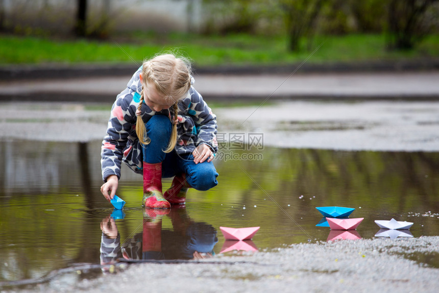 女孩经营粉红色纸船在雨中的水坑里玩耍图片