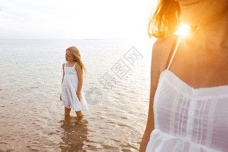 母亲和女儿穿着白裙子在日落时海边散步图片