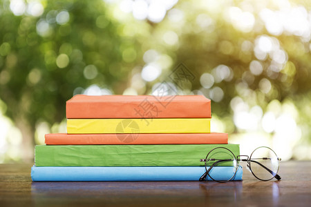 木桌上有眼镜的彩色叠书本有浅色背景的模糊树本图片