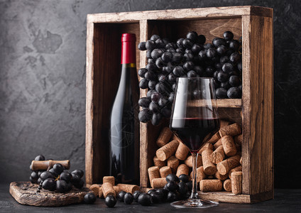红酒瓶装深葡萄和木箱的软黑石底的旧木箱内装着黑葡萄板上装着软木瓶的优雅葡萄酒杯图片