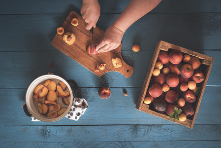 装有桃子和杏的木箱以及一名妇女用手在剪切板上和锅里在蓝桌上切水果图片