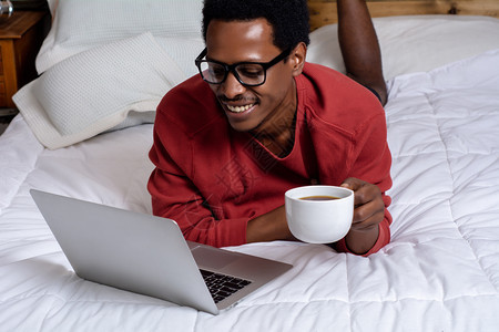年轻的美国男子使用笔记本电脑在床上喝咖啡图片