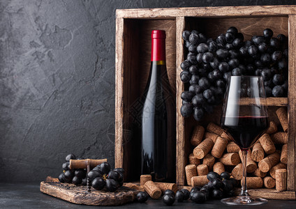 红酒瓶装深葡萄和木箱的软黑石底的旧木箱内装着黑葡萄板上装着软木瓶的优雅葡萄酒杯图片