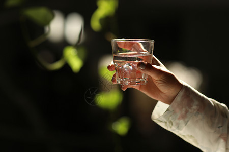 妇女双手被孤立用绿叶将一杯水握在黑暗背景上太阳光线落在玻璃上健康早晨图片