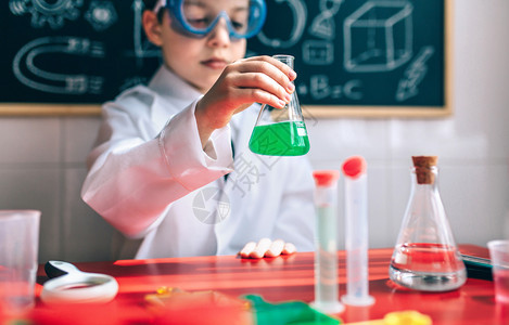小男孩科学家戴眼镜的男孩科学家拿着装有化绿色液体的瓶子反对抽黑板图片