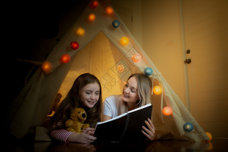快乐的小女孩和母亲在家中帐篷里一起微笑阅读书图片