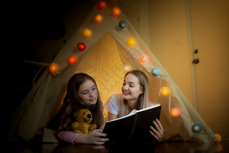 快乐的小女孩和母亲在家中帐篷里一起微笑阅读书图片
