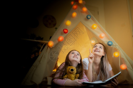 快乐的小女孩和母亲微笑着看书一起在家中帐篷指向天空创造节日思维概念图片