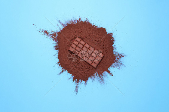 可粉堆和一根棕色巧克力条放在蓝色背景上放在整个巧克力和主要成分可粉堆上面图片