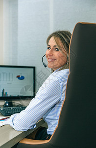 微笑着的金发秘书拿着耳机看相和办公室的电脑一起工作图片