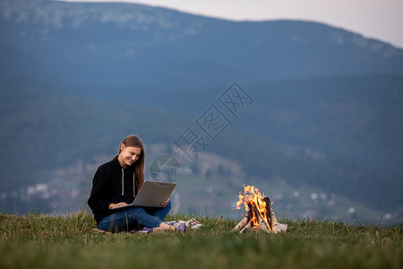 女孩坐在草地上工作营火在边上点燃工作商业自由职登记场所山区有笔记本的年轻女孩坐在草地上工作营火在边上点燃工作登记场所图片
