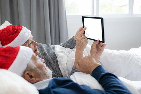 在卧室圣诞节日使用白屏幕背景的平板电脑上使用红帽子的男女老夫妇在卧室的圣诞节日使用白屏幕背景退休爱人家庭生活方式概念图片