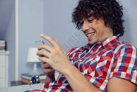 年轻英俊男人看着智能手机笑在家里的休闲时间概念上笑着图片
