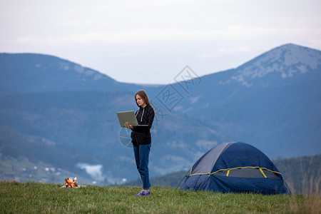 旅游女孩站在帐篷附近的一座山上吃着晚餐烧火拥有笔记本的年轻女背景美丽的山上旅游女孩站在帐篷附近的一座山上烧着火图片