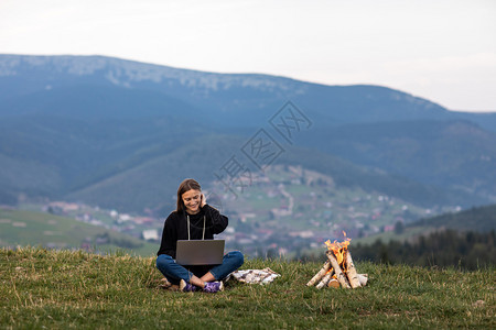 女孩坐在草地上工作营火在边上点燃工作商业自由职登记场所山区有笔记本的年轻女孩坐在草地上工作营火在边上点燃工作登记场所图片