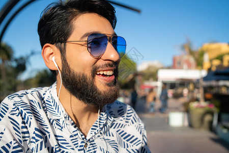 年轻英俊男人的肖像享受阳光灿烂的一天并用户外耳机聆听音乐城市概念图片