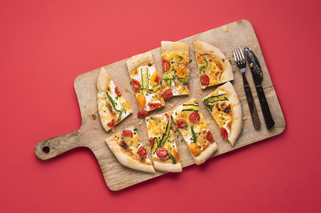 带新鲜蔬菜的春季比萨饼切片的在有餐具木板上红色背景的图片
