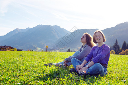 两个微笑的女孩坐在绿色草地上和山的背景图片