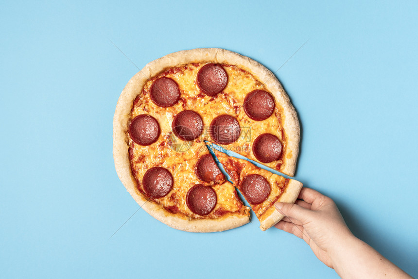 女人在蓝色背景上吃一块沙拉米比萨上面是辣椒披萨和一只手拿披萨快速食物美味图片