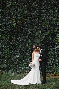新娘穿着长的白色礼服带着结婚花束和新郎一起穿着时装的新郎在前墙举行盛装婚礼后背景图片