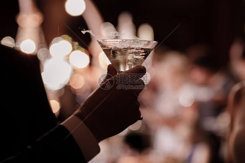 身着正式服装在派对背景上举着一杯马提尼的男子和身着马提尼杯的男子和手图片