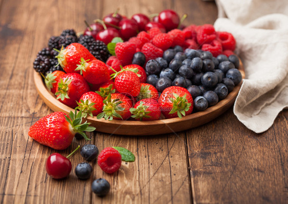 浅木质桌底的圆盘中新鲜的有机夏季果子混合物草莓蓝黑和樱桃并配有麻布厨房毛巾图片