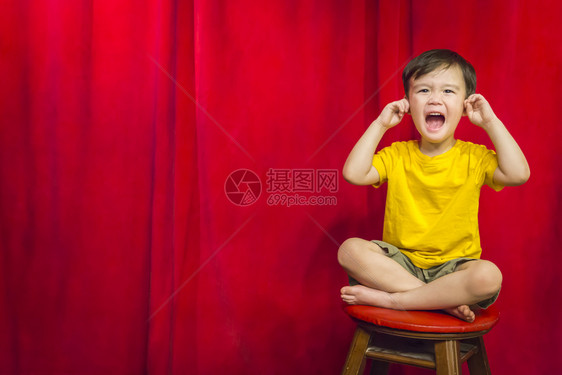 混血男孩他耳朵里有手指坐在红幕前的凳子上图片