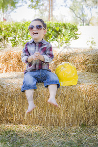 可爱的混合种族男孩笑着用太阳镜和硬帽外面坐在干草泥巴上图片