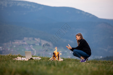 女青年自由职业者坐在火边晚上山暖他的手女旅游者在户外休息和笔记本电脑上工作女青年自由职业者坐在火边晚上山暖他的手女旅游者在户外用图片