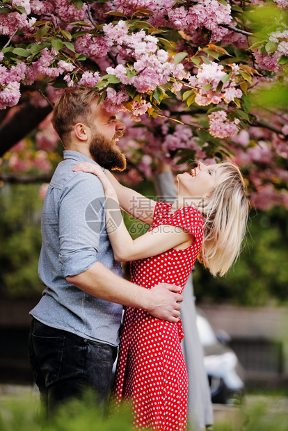 在萨库拉树附近的一对有时髦夫妇美丽年轻夫妇有胡子的男人和金发女郎在春天公园拥抱和欢乐概念的春天时尚和美丽图片