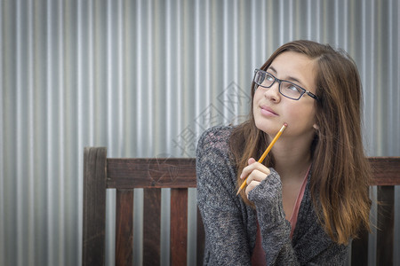 年轻漂亮的白日梦女学生用铅笔坐在长椅上看着一边图片