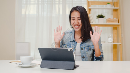 自由职业妇女随意使用平板电脑工作话视频会议与工作场所的客户在家中厅里举行快乐的年青轻女孩坐在办公桌上放松在互联网上做工作图片