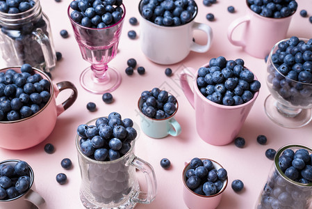 以不同大小和颜色的杯子和装有机蓝莓的夏季果实新鲜收获粉红色桌上的罐子有大量蓝莓图片