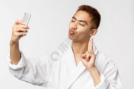 美spa和技术概念美穿着浴袍的傻亚洲男子闭上眼睛亲吻脸拍自在移动相机上展示和平标志站在白色背景美丽穿着浴袍的傻亚洲男子图片