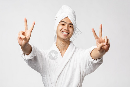 美丽spa和休闲概念穿着浴袍和巾的快乐英俊亚洲男子展示和平迹象并带着欢乐表达的热情站在白色背景的微笑美丽穿着浴袍和巾的快乐亚洲男图片