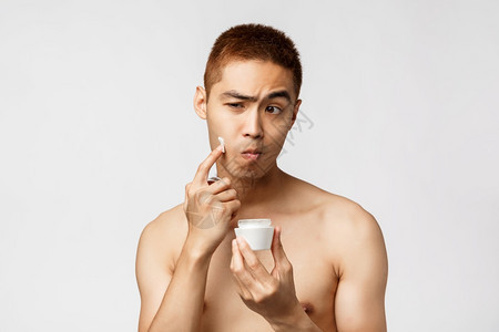 美丽人与休闲概念英俊的亚洲男子站在浴室里看着镜子运用日常的皮肤护理规奶油站着白色背景英俊的亚洲男子站在浴室里看着日常的皮肤护理规图片