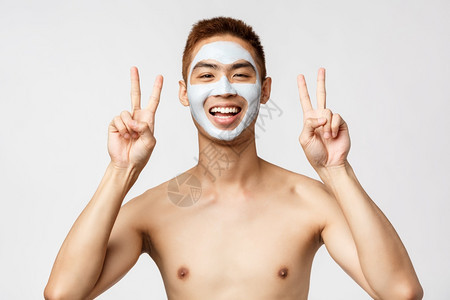 穿着躯的快乐年轻亚洲男子运用皮肤常规化妆品面罩展示和平标志在家放松自我持白背景美丽皮肤护理和spa概念穿着躯的快乐年轻亚洲男子运图片