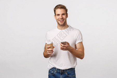 愉快的现代金发男子手持纸杯咖啡喝茶手持智能机发短信微笑的相机浏览互联网在休息期间网上购物图片