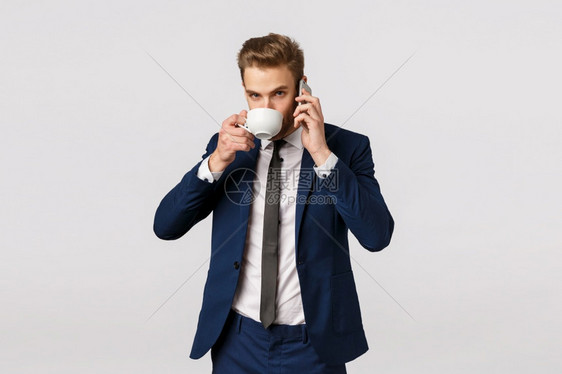 没有时间休息工作第一具有吸引力的年轻商人穿经典西装在电话交谈时喝杯子咖啡把智能手机贴近耳朵打电话助理拿洗衣服没有时间休息在电话交图片