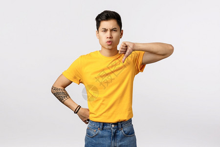 你看起来像个失败者不高兴和失望的年轻英俊亚洲男子纹着黄色t恤衫牛仔裤展示拇指低缩和假人怀疑判断坏的玩耍不赞成你看起来像失败者不满图片