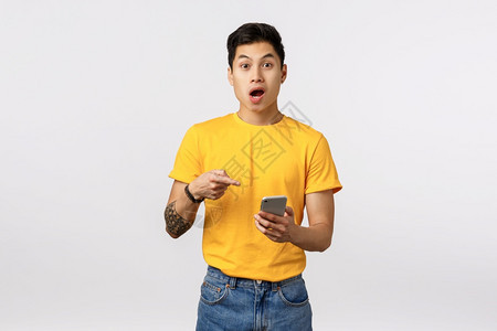 使用黄色t恤衫持有智能手机对着显示和喘着迷令人惊讶热的亚洲人穿黄色t恤衫握着智能电话对手机显示和喘迷震惊用过滤器应程序白色背景编图片