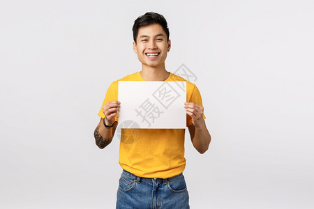 友善流出和快乐的年轻亚洲男子身穿黄色t恤衫持有空白的纸片笑得过快写口供或陈述展示公司宣传白色背景友好流出和快乐的年轻亚洲男子身穿图片