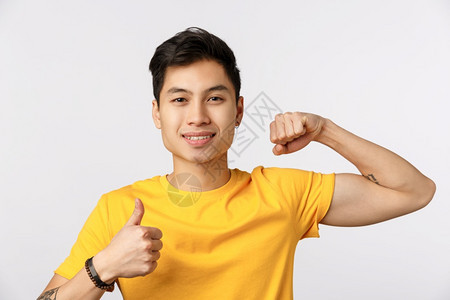 穿着黄色t恤的有吸引力年青亚洲男子穿着黄色t恤展示肌肉和拇指微笑鼓励体操锻炼一起健身想要获得强壮的biceps时间泵起来穿着黄色图片