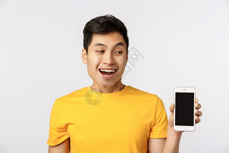 喜欢新过滤器应用程序喜欢黄色t恤衫的有吸引力亚洲男子显示智能手机看着移动屏幕笑着的娱乐喜欢服务如何真棒的功能喜欢新的过滤器应用程图片