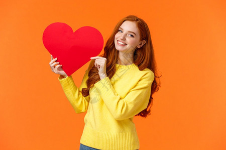 情人节是供认的完美时机穿着黄色毛衣的可爱浪漫和温柔的红发女人拿着大胸牌微笑表达感情和浪漫站立橙色背景穿着黄色毛衣的可爱浪漫和红发图片